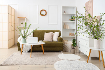 Green sofa, coffee table and vase with blossoming branches in light living room