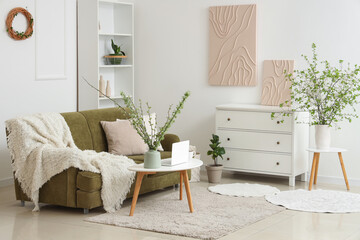 Green sofa, coffee table, chest of drawers and vase with blossoming branches in light living room