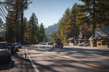 Sunny day on mountain street lined with coniferous trees, vehicles parked, few cars driving. Red...
