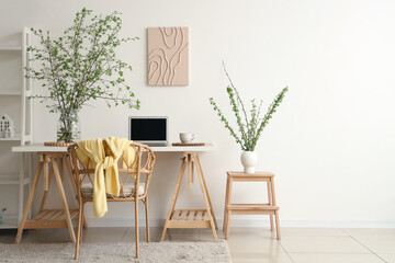 Stylish workplace, armchair, laptop and vase with blossoming branches in light living room