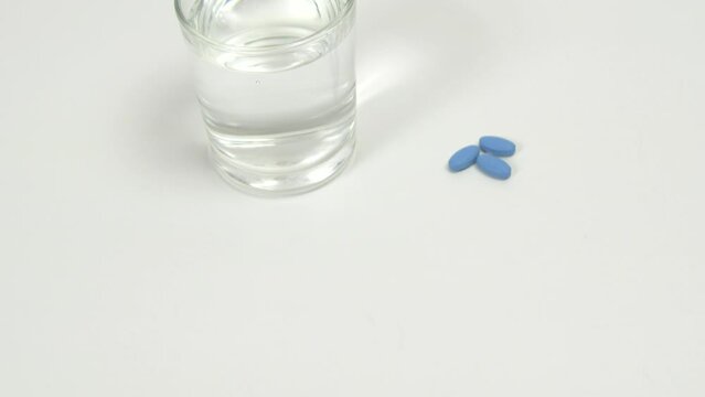 Blue pills for treating erectile dysfunction on white table with glass of water. Pan up
