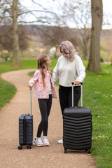 Woman and girl having a conversation while walking with suitcases in a park.