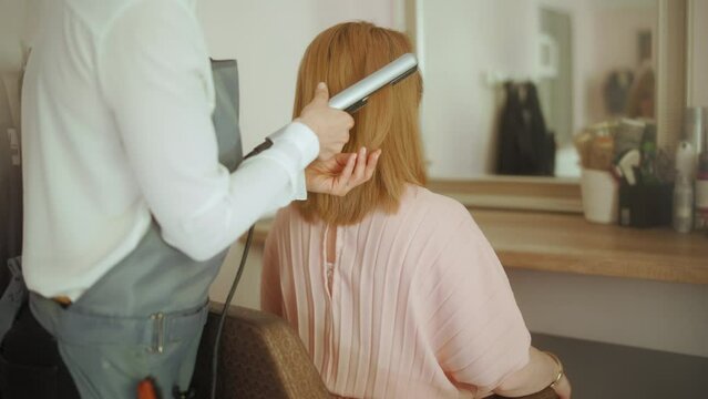 Seen from behind hair salon employee in modern beauty studio with hair straightener and client.