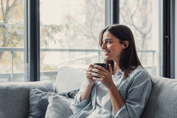 Cheerful dreamy girl holding mug of hot beverage, looking away in deep good thoughts, smiling,...