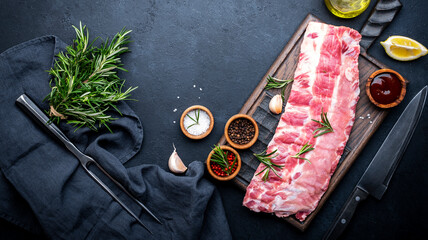 Raw pork ribs with rosemary, spices and barbecue sauce on oak wooden cutting board prepared for cooking on black kitchen table background, top view