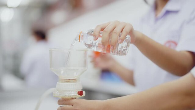 A woman is holding a glass beaker and a bottle of liquid. The scene is set in a laboratory, and the woman is a scientist or a lab technician. The atmosphere of the image is focused and professional