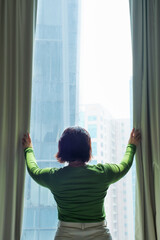 A woman opens curtains and looks out window.