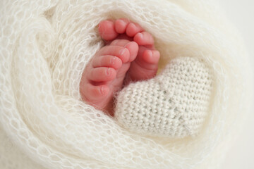 The tiny foot of a newborn baby. Soft feet of a new born in a white wool blanket. Close up of toes, heels and feet of a newborn. Knitted white heart in the legs of a baby. Macro photography