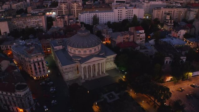 4k drone footage. Camera flies up revealing a view of the Romanian Athenaeu, a concert hall in the center of Bucharest. One of the main landmarks of the Romanian capital. Evening city lights at dusk