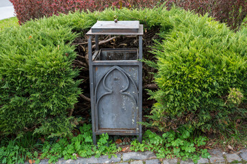 Gray metal trash can in ornamental green bushes.