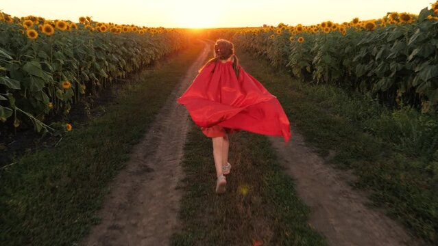 Cute teen girl superhero running in red cloak fantasy imagination at sunflower field sunset back view. Female kid child hero image flying pretend powerful character planet protection at flower meadow