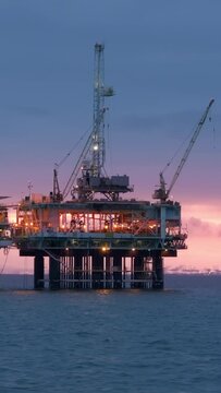 Vertical video. An oil rig stands tall amidst the vast expanse of the ocean, with pipes and machinery extending outwards. The rig is actively extracting oil, with workers visible on its structure.