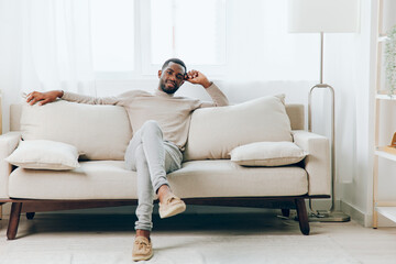 Sad African American man sitting on a comfortable sofa in his modern living room, lost in thought...