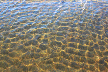 Close-up of a river bed with yellow sand, stones curling from the wave, in bright sunlight with beautiful highlights on the water. Texture of river sand. Texture with seashells and pebbles on the wet 