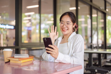 A cheerful female student sits at a table in a cafeteria talking on a video call with her friend.