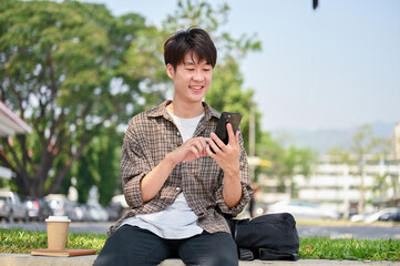 A cheerful young Asian male college student sits on a stone bench in a park, using his smartphone.
