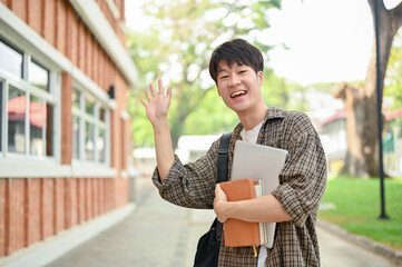 A friendly Asian male student stands outdoors on campus, waving his hand and smiling at the camera.
