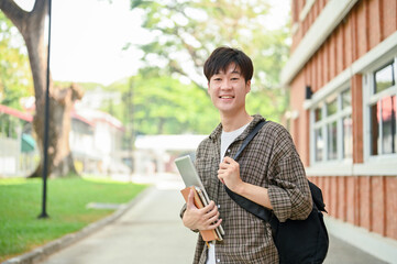 A cheerful Asian man student stands outside a brick building on campus, holding a laptop and books.