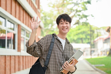 A friendly Asian male student stands outdoors on campus, waving his hand and smiling at the camera.