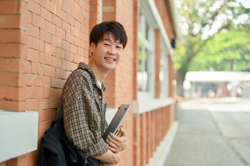A smiling young Asian male college student stands by a brick wall on campus, carrying a backpack.
