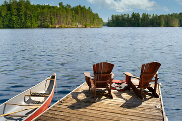 Two Adirondack chairs on a wooden dock overlook the calm lake. Life jackets are visible near the chairs. A red canoe is tied nearby. Across the water, cottages are visible. - Powered by Adobe