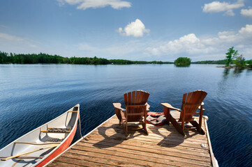 Two Adirondack chairs on a wooden dock overlook the blue waters of a Muskoka lake in Ontario,...