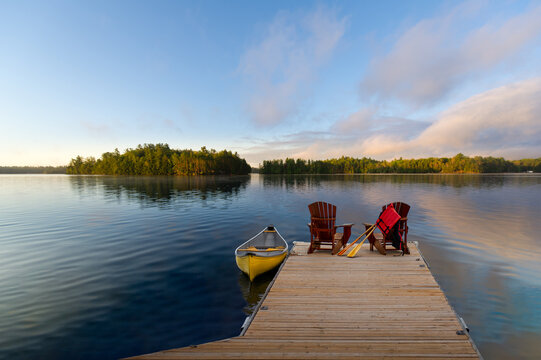 Two Adirondack chairs on a wooden dock overlook the calm lake. A life jacket and paddles hang from one chair, while a vibrant yellow canoe is tied nearby. Across the water, cottages are visible. - Powered by Adobe
