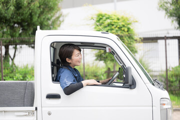 新緑の中トラックを運転する中年の作業着の女性の横顔　運送や配送業などのイメージ　廃品回収