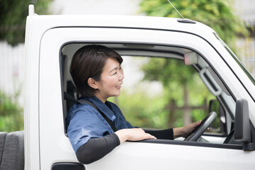新緑の中トラックを運転する中年の作業着の女性の横顔　運送や配送業などのイメージ