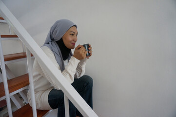 Asian muslim woman sits on stairs, drinks hot tea and enjoying peaceful. Leisure time at home...