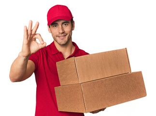 Young Hispanic delivery man with boxes cheerful and confident showing ok gesture.