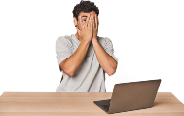 Hispanic young man working on laptop blink through fingers frightened and nervous.