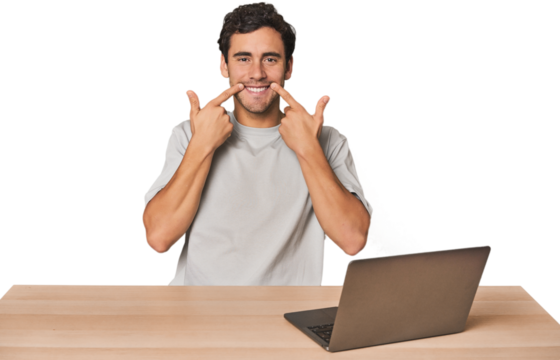 Hispanic young man working on laptop smiles, pointing fingers at mouth.