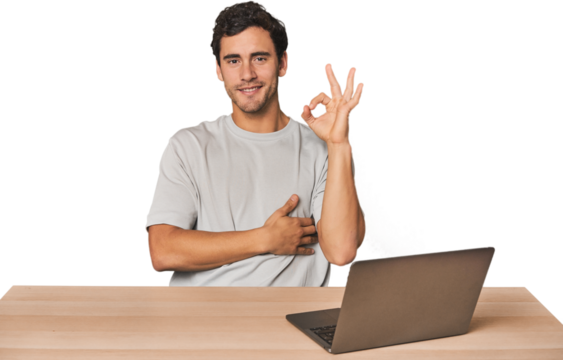 Hispanic young man working on laptop winks an eye and holds an okay gesture with hand.