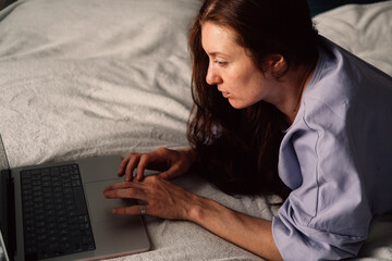 A woman lies on her bed deeply focused on her laptop screen