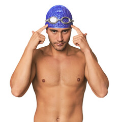 Young Hispanic man with swim gear focused on a task, keeping forefingers pointing head.