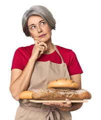 Caucasian mid-age baker with bread loaves looking sideways with doubtful and skeptical expression.