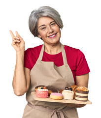 Caucasian mid-age baker with pastry tray joyful and carefree showing a peace symbol with fingers.
