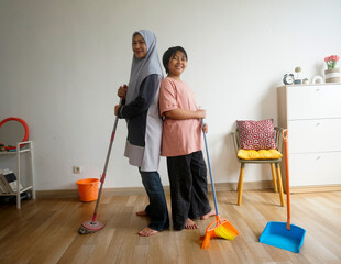 Happy Asian muslim mother and daughter standing back to back while cleaning house