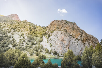 Mountain panorama, landscape with rocky peaks and blue turquoise lake Chukurak in the Fan Mountains in Tajikistan, hills covered with forest on a sunny summer day