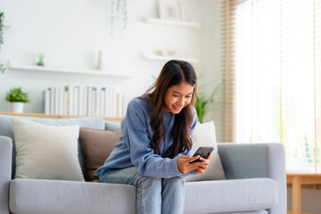 Happy young asian woman relax on comfortable couch at home texting messaging on smartphone, smiling girl use cellphone, chatting online message, shopping online from home