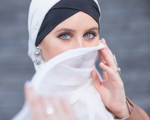 Portrait of a young blue-eyed woman in a hijab against a gray brick wall. Muslim woman with white...