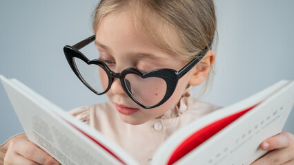 Portrait of a little girl in heart-shaped glasses reading a book. 