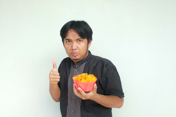 A young Asian man with a happy expression holds a bowl of potato snacks with thumbs up gesture....