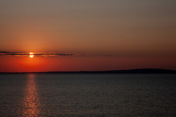 Sunset over the sea against the background of the coast. Evening on the sea calm in clear weather. The disk of the sun in the evening sky over the sea.