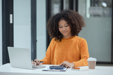 African American female working with laptop and calculator at desk in office.