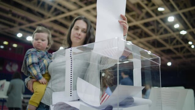 Close up shot of polling box standing on table in polling station. American female voter with kid on hands puts bulletin into box. Political races of US presidential candidates. National Election Day.