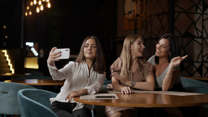 Girlfriends take a selfie on a mobile phone camera while sitting in a cafe. Three attractive girls...