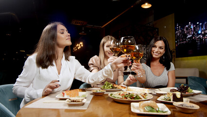 Three cute girlfriends having a rest sitting at a table and drinking wine merrily talking. Girls clink glasses with wine glasses.