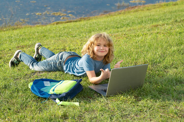 Child lying on lawn in the park on beautiful summer day, doing homework on laptop.